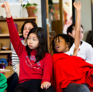 two kids in a classroom raising their hands